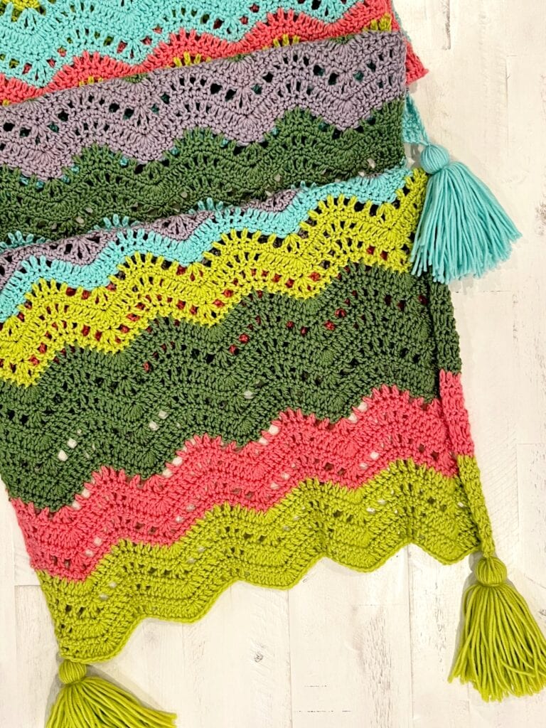 A colorful crocheted blanket featuring a chevron zigzag pattern in green, yellow, pink, purple, and blue—this cozy throw is inspired by a Crochet Chevron Wrap Pattern and finished with large tassels at each corner. Close-up of crochet chevron stitches showing peaks, valleys, and color changes. -Marly Bird