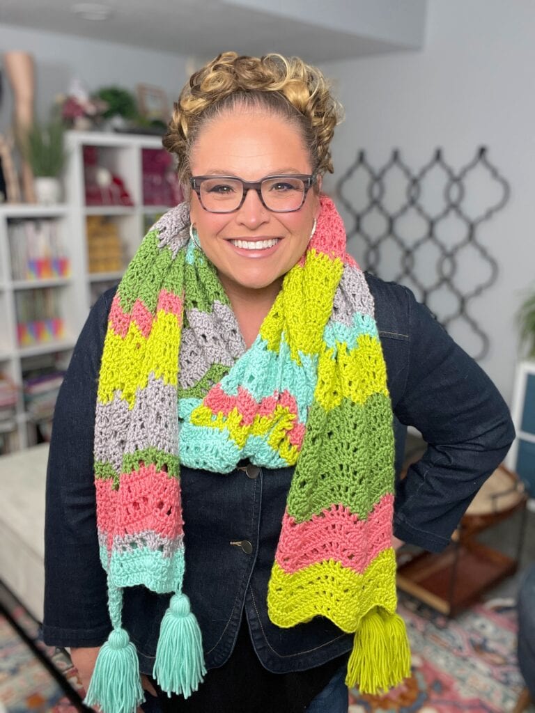 A smiling woman with curly blonde hair and glasses wears a colorful crochet chevron wrap with tassels. She stands indoors in a cozy, well-lit room filled with shelves, books, and plants—perfect for showing off this free crochet pattern by Marly Bird. -Marly Bird