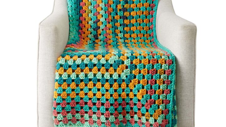 A Tunisian crochet blanket with bright squares and large teal tassels draped over a cream armchair, showing clear stitch detail.