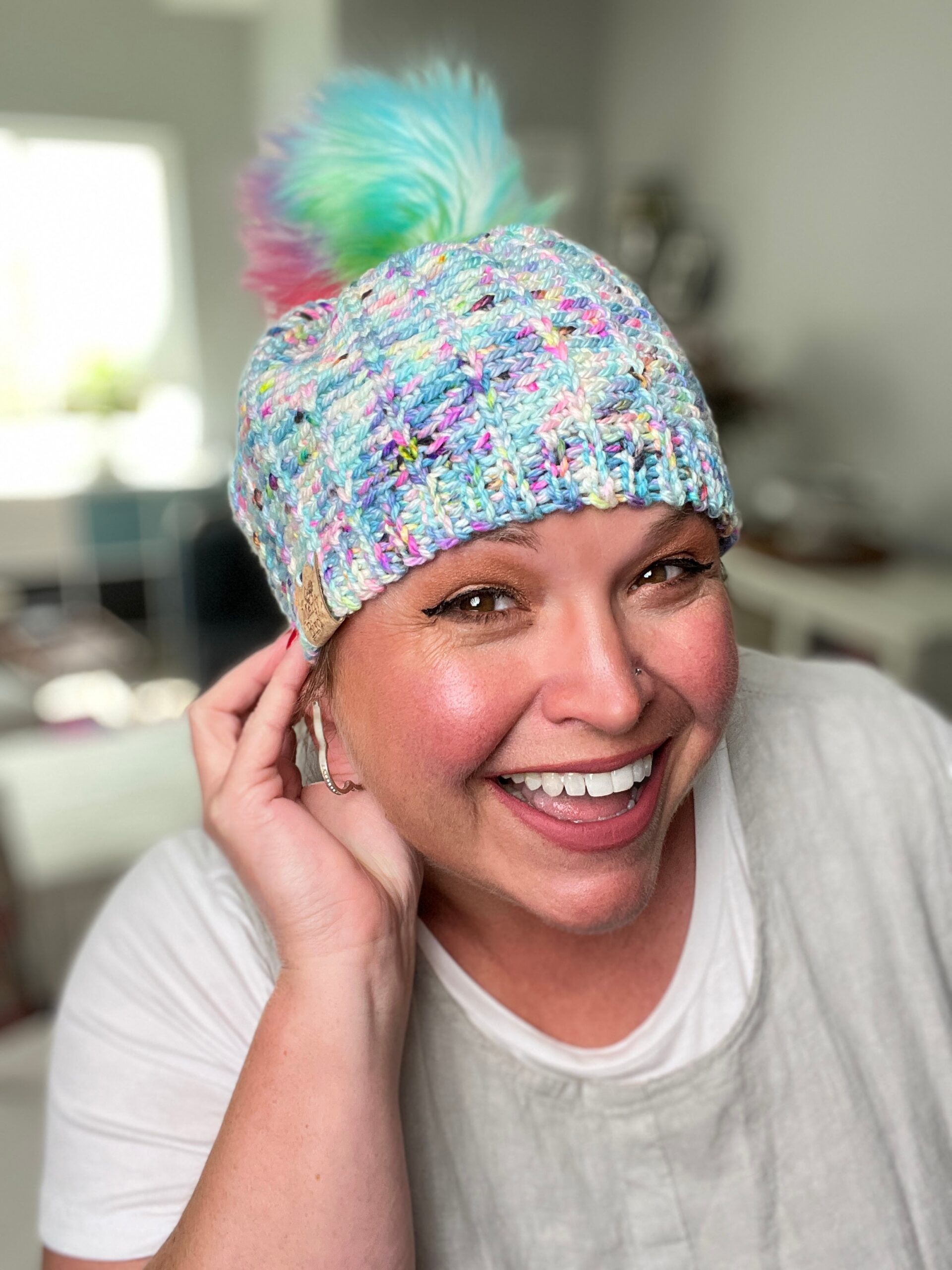 A person smiling broadly, wearing a colorful knit-look crochet hat with a pastel pom-pom. They are indoors with a blurred background that includes furniture and bright light from a window. The person is touching the hat with one hand and has a happy expression. -Marly Bird