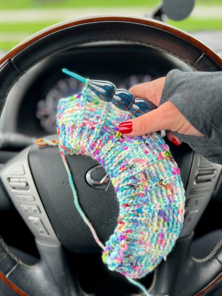 A person with red nail polish and a gray sleeve is holding colorful knitting on circular needles over a steering wheel inside a car, perfecting that knit-look. The wooden trim wheel features a logo with a silver 'L' in the center while the background is blurred. -Marly Bird