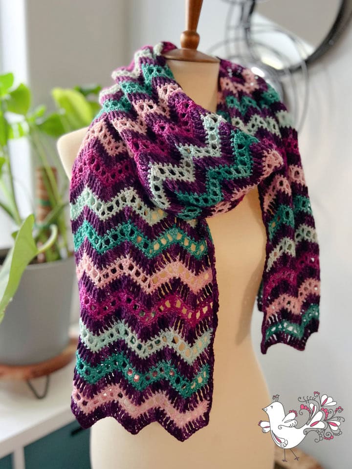 A mannequin displays a crocheted scarf with a chevron pattern in purple, teal, pink, and white. A potted plant and round mirror add charm to the scene, perfect inspiration for those seeking free knitting and crochet patterns. A small bird design decorates the corner. -Marly Bird