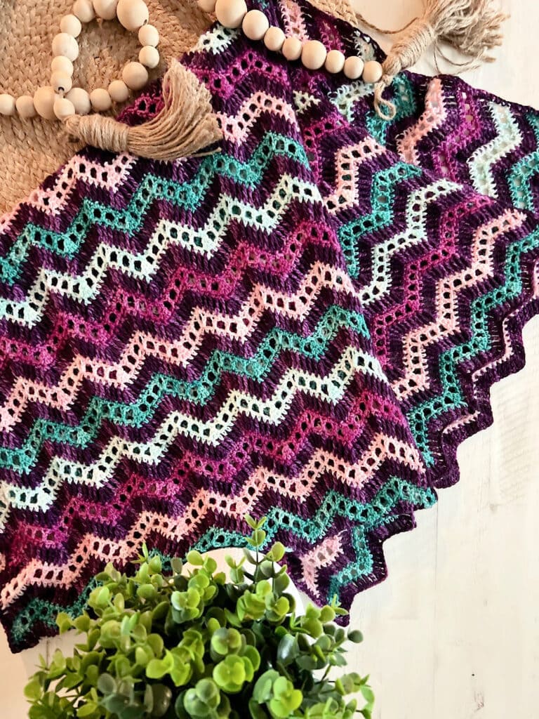 A colorful crocheted zigzag-patterned blanket in shades of purple, pink, teal, and cream is spread out on a white surface, with a plant and wooden-bead tassel decoration nearby—perfect inspiration for free knitting and crochet patterns. -Marly Bird