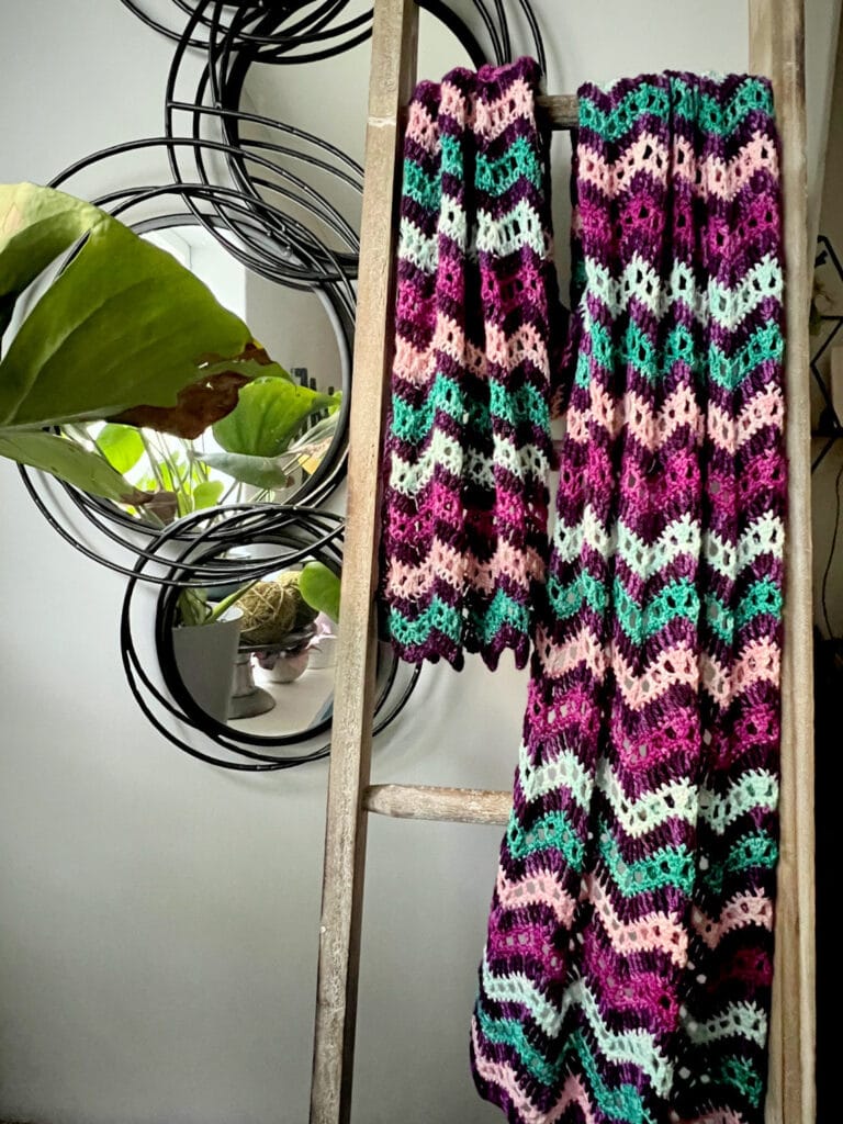 A colorful crocheted blanket with purple, pink, green, and black stripes hangs on a wooden ladder. A plant and decorative round mirrors are in the background—perfect for fans of free knitting and crochet patterns. -Marly Bird