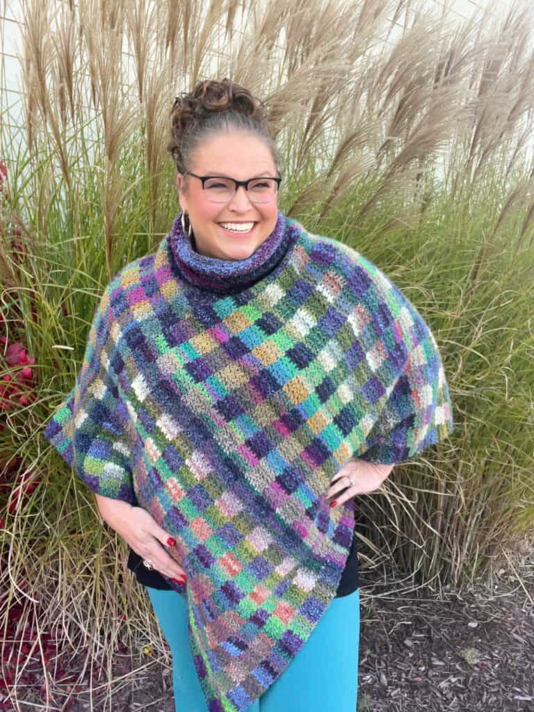 Marly Bird smiles while standing in front of tall grasses. They wear glasses, a crochet poncho reminiscent of Marly Bird's designs, and turquoise pants. Their hair is styled up, and they have hoop earrings. -Marly Bird