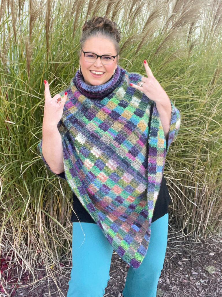 A smiling woman standing in front of tall grass, wearing glasses and a multicolored crocheted poncho with a cowl neck, playfully gesturing the 'rock on' sign with both hands. - Marly Bird - Check Me Out Crochet Poncho