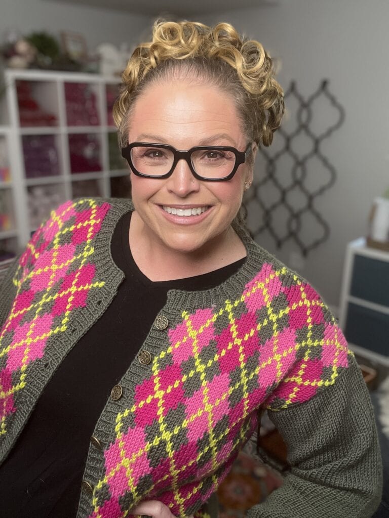 A smiling woman with curly blonde hair and black glasses wears a green cardigan with a pink and yellow argyle pattern, standing in a cozy room with shelves, patterned decor, and a Dream Intarsia Knit Blanket in the background. -Marly Bird