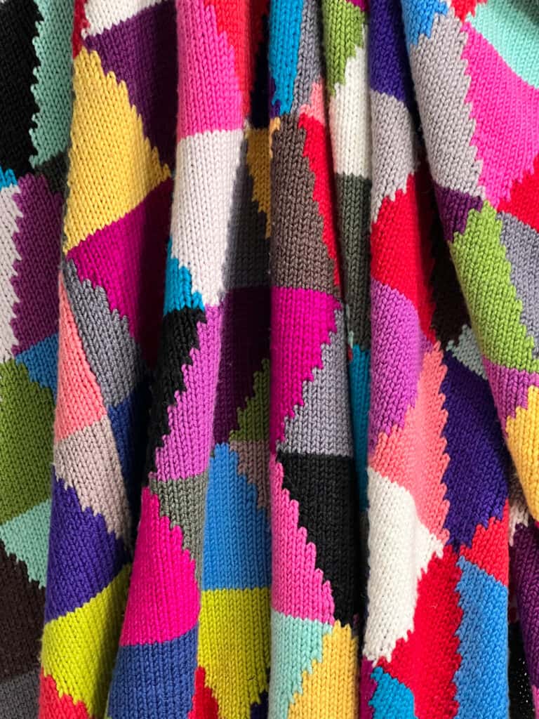 A colorful knitted fabric reminiscent of the Chic Sheep Dream Intarsia Knit Blanket, with a patchwork pattern featuring vibrant colors like red, blue, yellow, pink, and purple in a geometric zigzag design. The texture and folds of the fabric are beautifully visible. -Marly Bird