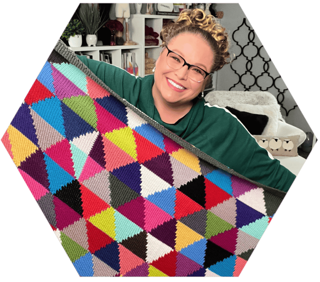 A person with glasses smiles while holding up a vibrant, multicolored quilt reminiscent of the Chic Sheep Dream Intarsia Knit Blanket. The background features shelves adorned with books and decor. -Marly Bird