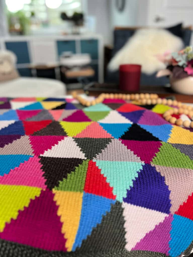 A colorful quilt featuring a chic pattern of multicolored triangles is displayed on a table. In the blurred background, there are shelves, a fluffy cushion, and some decorative items. The overall setting is cozy and dream-like, perfect for showcasing an intarsia knit blanket. -Marly Bird