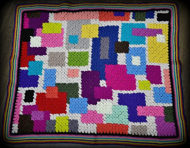 A colorful FREE Crochet Chic Sheep Blanket featuring irregular, blocky patches in vibrant pink, blue, white, yellow, green, and purple hues, all finished with a striking striped multicolor border. -Marly Bird