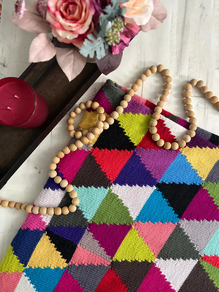 A colorful, geometric-patterned textile, inspired by free knitting and crochet patterns, features small wooden beads draped on top, next to a tray holding a pink candle and a bouquet of artificial flowers on a white wooden surface. -Marly Bird