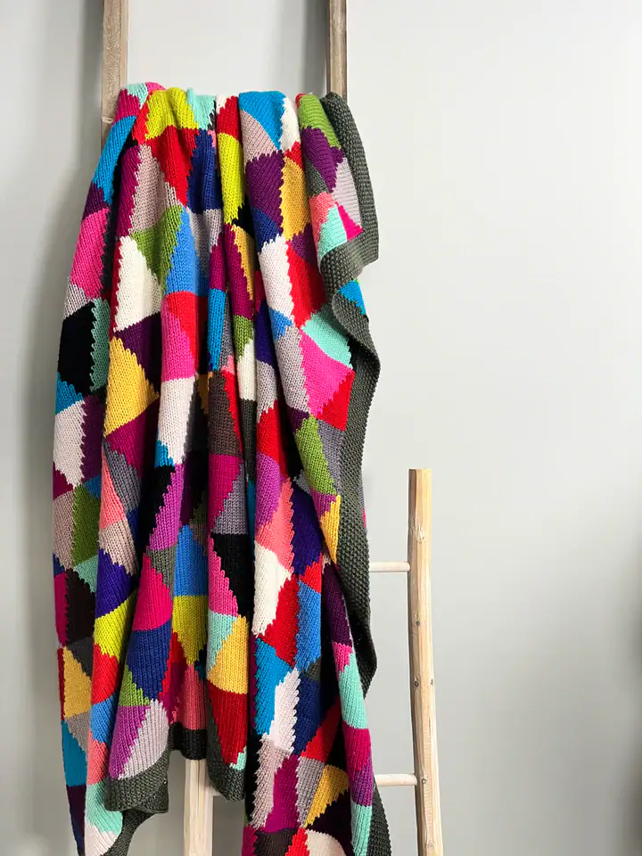 A colorful, geometric patchwork knit blanket—crafted from free knitting and crochet patterns—features bright triangles and drapes over a light wooden ladder against a plain light gray wall. -Marly Bird