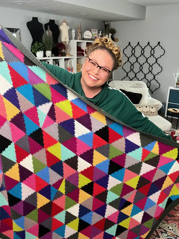 A smiling person with glasses holds up a colorful, geometric patchwork quilt in a cozy room filled with shelves, decorations, crafts, and free knitting and crochet patterns in the background. -Marly Bird