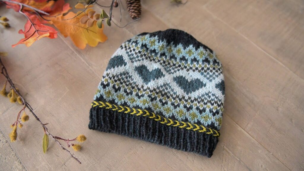 A stranded knit Hat with a Fair Isle pattern featuring hearts and diamonds in various shades of blue, grey, and black. The beanie has a yellow decorative braid near the base. It is displayed on a wooden surface, accompanied by autumn-themed foliage. Pattern available. -Marly Bird
