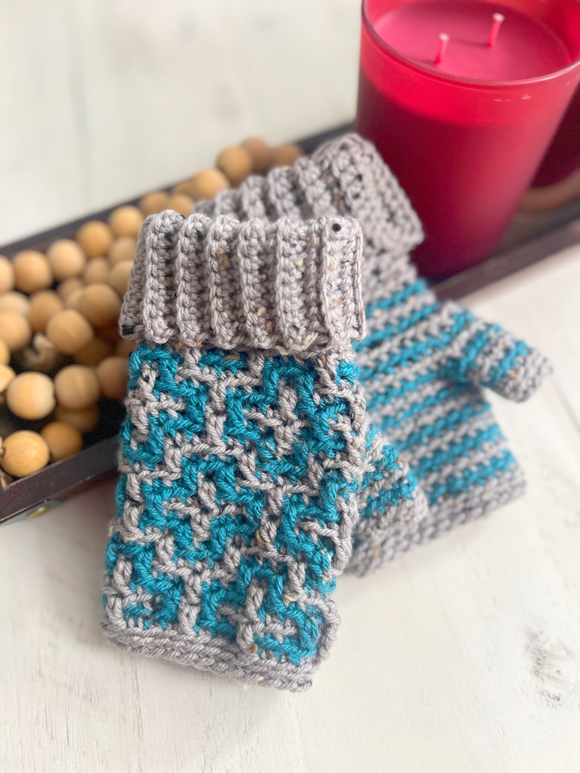 A pair of knitted fingerless gloves with intricate patterns in gray and teal are placed on a table, exuding cozy Cinnamon Dreams vibes. Next to them is a red candle in a glass jar, adding warmth to the scene. The table decoration includes a tray with wooden beads, creating an inviting atmosphere. -Marly Bird