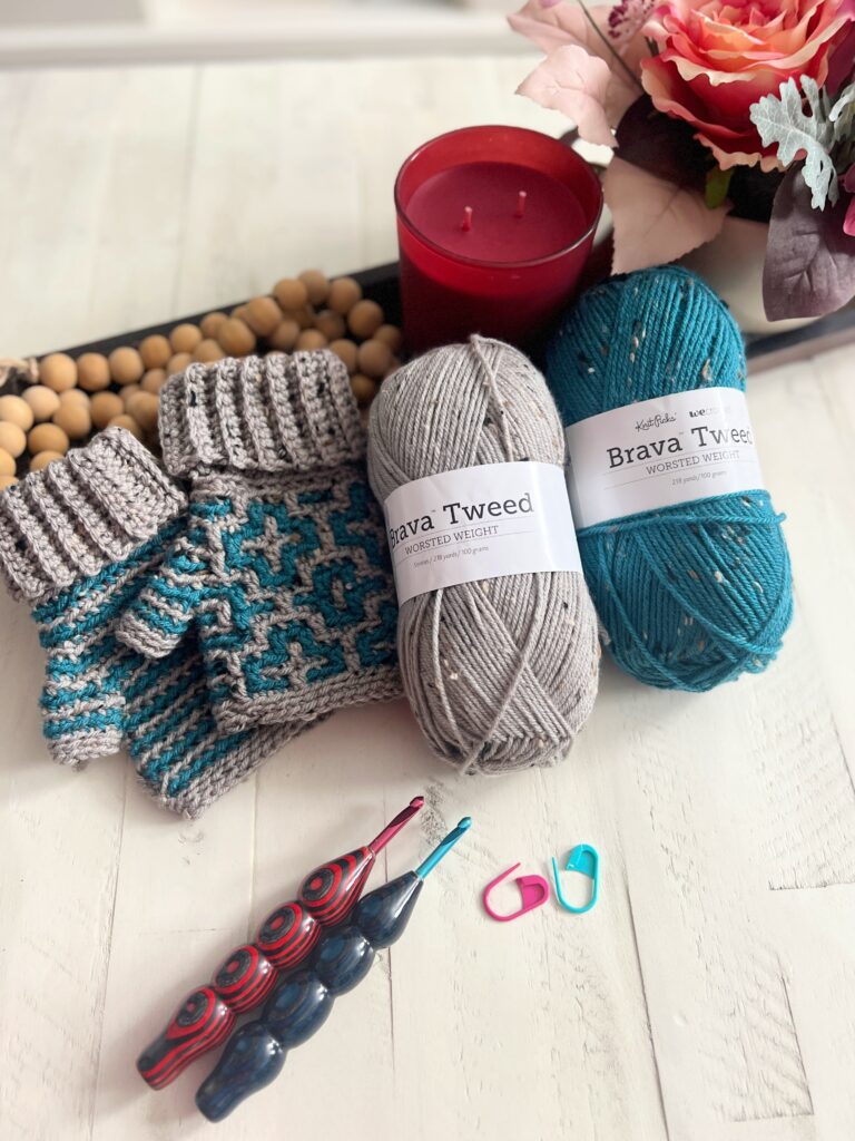 On the table, alongside two balls of Brava Tweed yarn in gray and teal, lie knitted fingerless gloves, vibrant crochet hooks, stitch markers, a red candle, and a decorative floral arrangement. A "Cinnamon Dreams" free pattern might inspire your next project. -Marly Bird