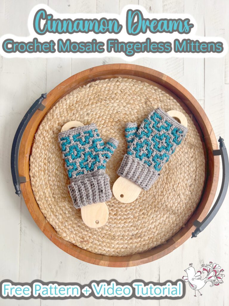 Displayed on wooden holders atop a round woven tray, the Crochet Fingerless Mittens showcase intricate blue and gray geometric patterns. The text reads "Cinnamon Dreams: Free Pattern + Video Tutorial," inviting you to create your own cozy masterpiece. -Marly Bird