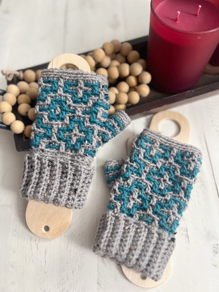 Gray and teal crochet fingerless mittens rest elegantly on wooden boards, accompanied by a red candle and wooden beads on a white surface. The charming scene whispers of Cinnamon Dreams, inviting cozy moments. -Marly Bird