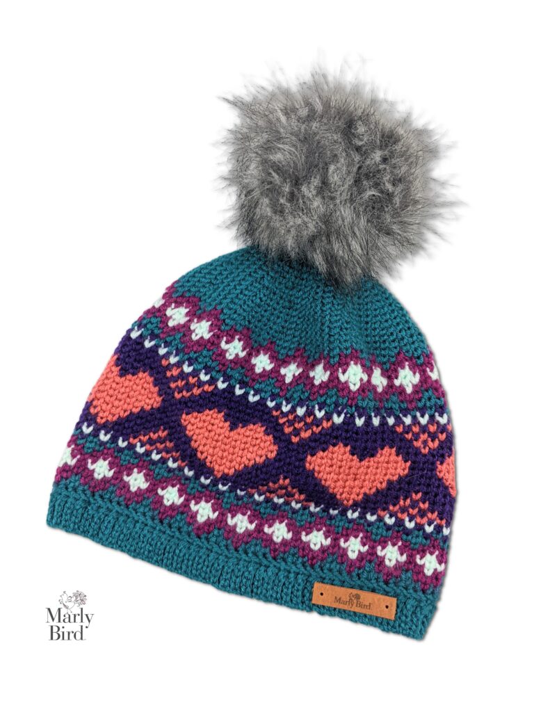 A knit-look crochet hat with a multicolored pattern, featuring designs of hearts and geometric shapes. The beanie has a teal base color, with pink, white, and purple accents. Topped with a fluffy gray pom-pom, it includes the Marly Bird logo on the bottom left. Find the free pattern online! -Marly Bird