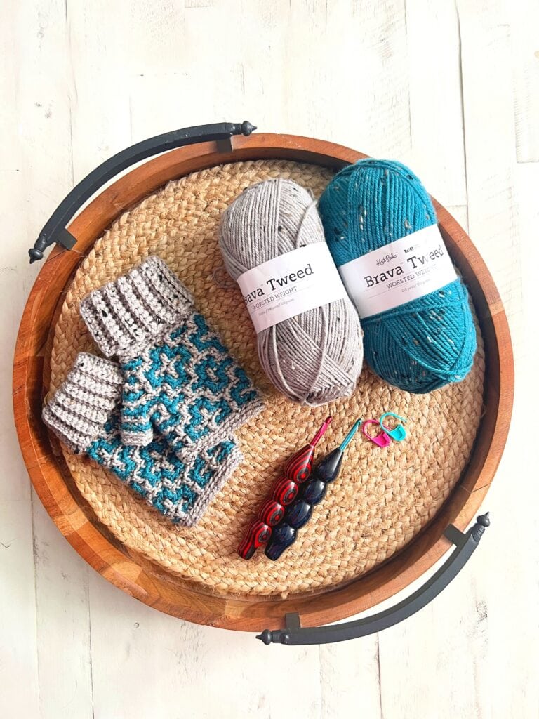 Fingerless gloves in blue and gray yarn rest on a tray with hooks, stitch markers, skeins, and a crochet and knitting pattern booklet.