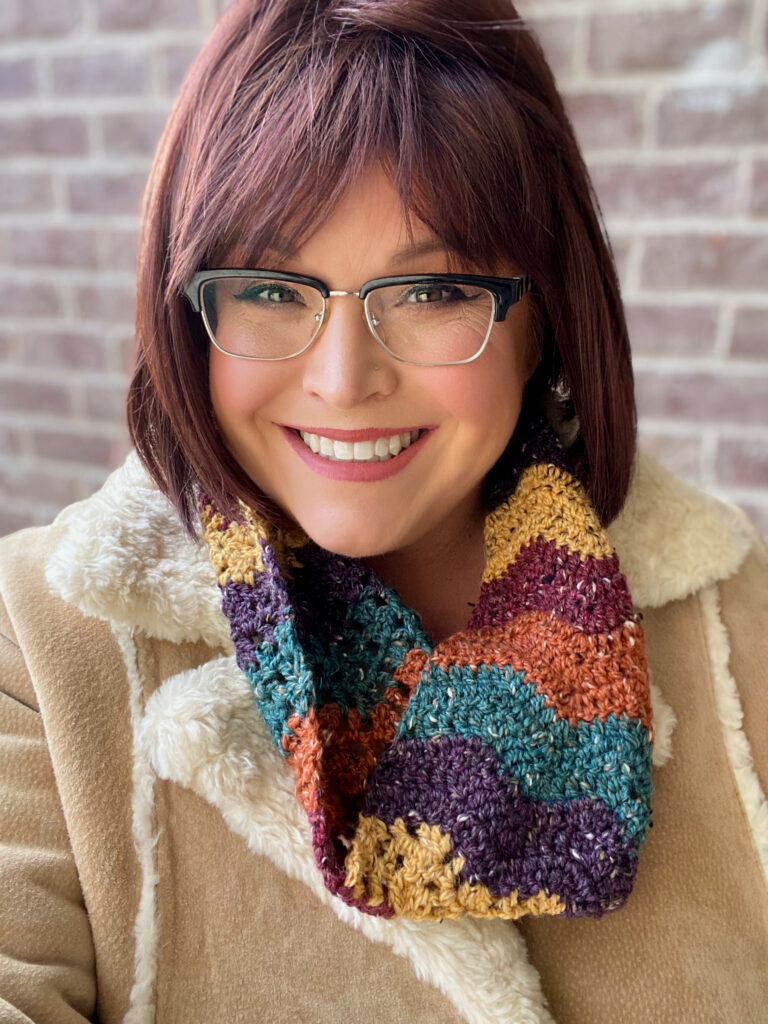 A smiling woman with short reddish-brown hair and glasses, wearing a colorful crochet-patterned scarf and a beige coat, standing in front of a brick wall. -Marly Bird