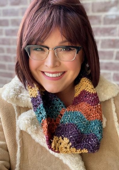 Marly Bird wearing City Tweed Chevron Cowl over light brown sheepskin coat, with brick wall background. Cowl crocheted in ripple stitch pattern in purple, teal, rust, wine, mustard colored yarn.