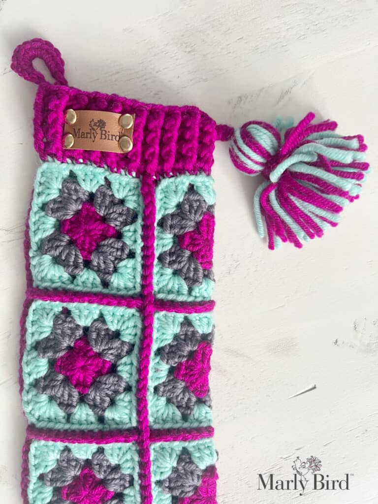 A crocheted pouch with a patchwork of colorful granny squares in pink, teal, and gray features a tassel and a small metal tag labeled "Marly Bird" on top. The background is a white surface with the "Marly Bird" logo—a delightful nod to traditional crochet stocking patterns. -Marly Bird