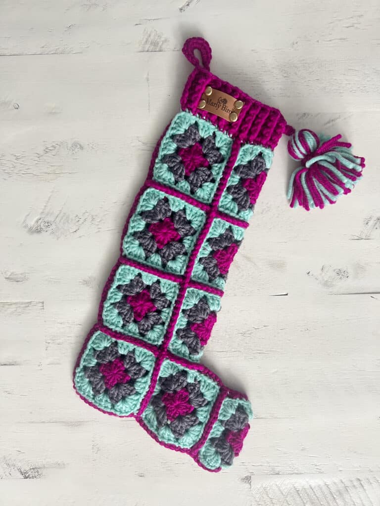 This crocheted Christmas stocking showcases a vibrant patchwork of granny squares in purple, turquoise, and gray. Complete with a decorative tassel and hanging hook, this stocking is perfect for adding charm to your holiday décor. Explore free patterns to create your own festive masterpiece! -Marly Bird