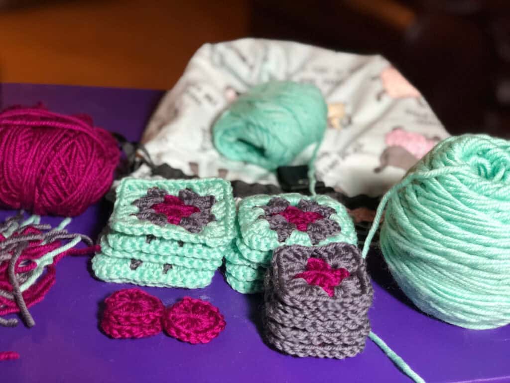 A close-up of classic crocheted granny squares in teal, gray, and burgundy yarn. Multiple skeins rest on a purple surface beside two small round crochet pieces and a yarn bag in the background, evoking a cozy stocking pattern waiting to be created. -Marly Bird