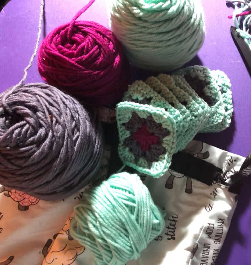 Colorful balls of yarn in pink, purple, gray, and mint green are scattered on a purple surface alongside a partially crocheted classic granny square piece with a floral pattern. A printed fabric bag with black drawstrings is nearby. -Marly Bird