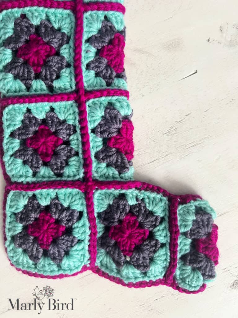 A crocheted Christmas stocking features colorful granny squares in shades of teal, gray, and magenta, creating a delightful patchwork pattern. With the Marly Bird logo in the bottom corner, this crochet treasure is perfect for those seeking a free granny square stocking pattern. -Marly Bird