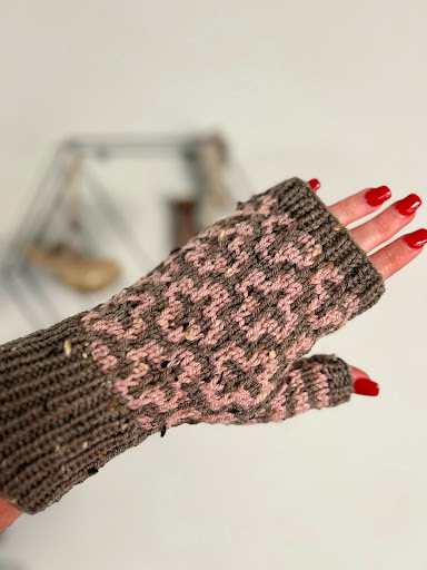 A hand adorned with a Cocoa Dreams fingerless mitten featuring a pink knitted pattern. The nails are painted red, while the softly blurred background showcases a geometric hanging decoration. -Marly Bird