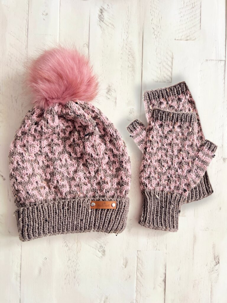 A pink and gray mosaic knit beanie with a pom-pom and matching fingerless mittens adorned in leopard print rests elegantly on a white wooden surface, capturing the essence of Cocoa Dreams. -Marly Bird