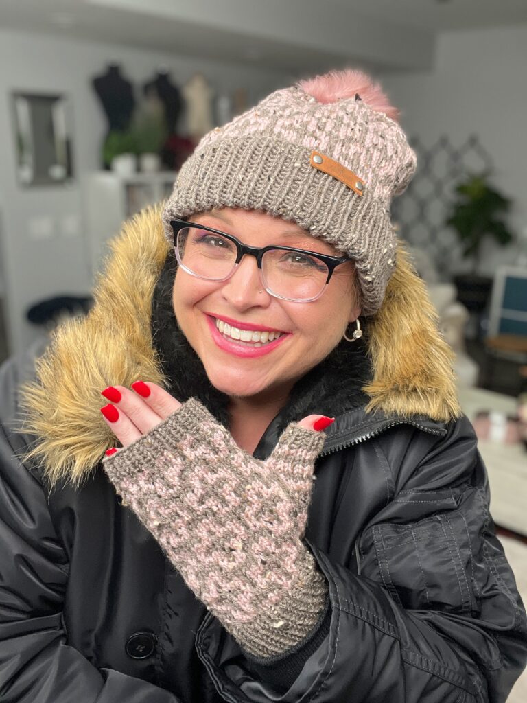 A person smiling warmly, wearing a pink and gray knitted hat with a pom-pom, glasses, and matching fingerless mittens made from a free pattern. They are also wearing a black coat with a fur-lined hood, embodying cozy Cocoa Dreams. -Marly Bird