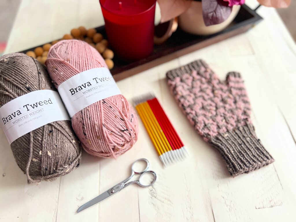 On a white surface, balls of brown and pink Brava Tweed yarn await beside small scissors, red and yellow knitting needles, and a pink knitted glove with a grey cuff. In the background, a red candle flickers atop a decorative tray. Dive into the Cocoa Dreams free pattern for cozy fingerless mittens. -Marly Bird