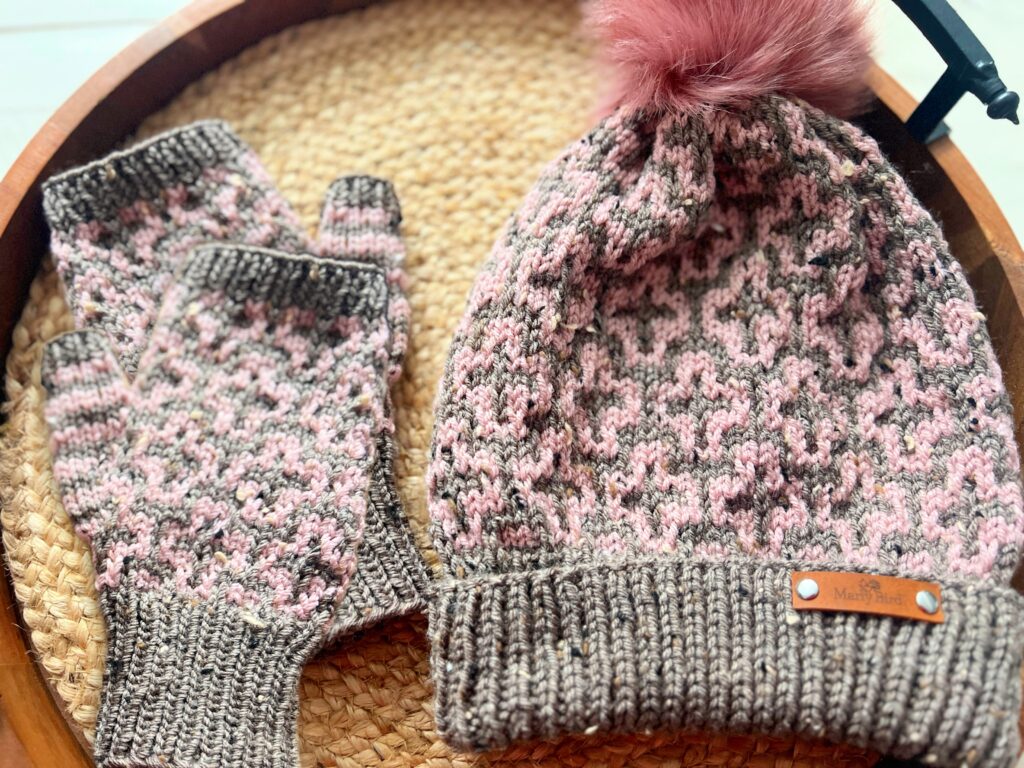 A knitted hat with a pink pom-pom and matching Cocoa Dreams fingerless gloves, featuring a pink and gray pattern, snugly placed on a woven basket. -Marly Bird