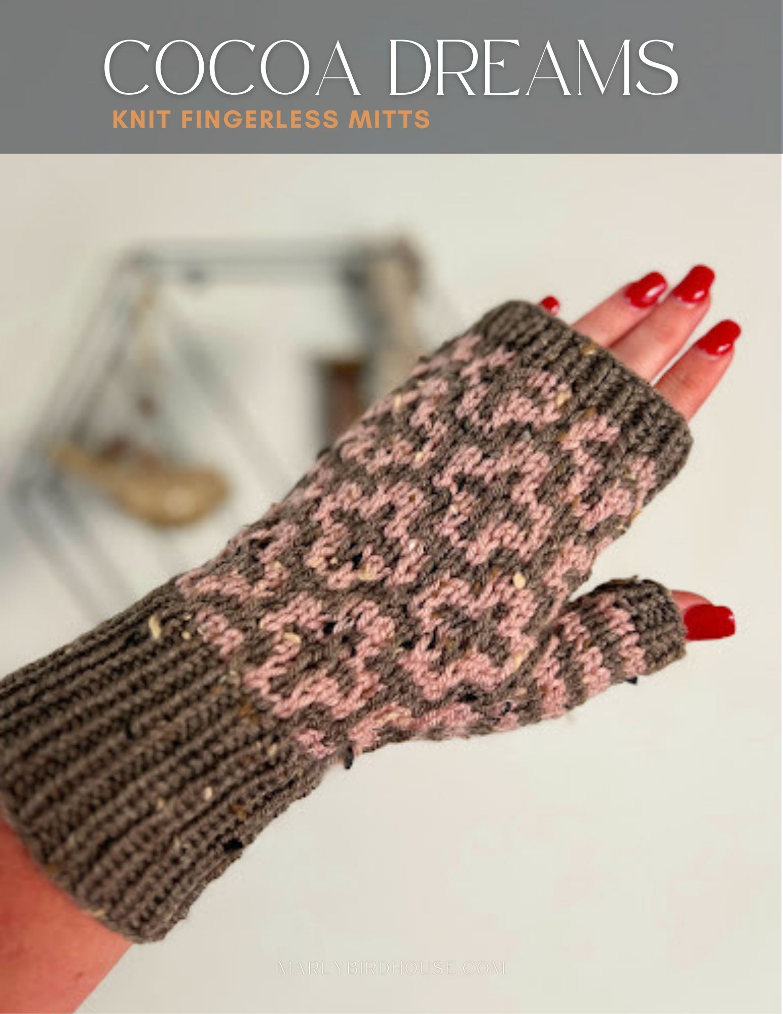 A hand displays a knit fingerless mitt with a brown and pink pattern, perfectly complementing its red-painted nails. The "Cocoa Dreams Knit Fingerless Mittens" stand out beautifully against a backdrop of blurred decorative art, offering both style and charm. -Marly Bird