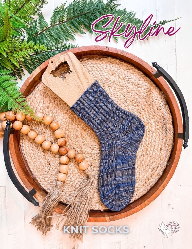 A blue and gray Ribbed Knit Sock is displayed on a wooden sock blocker atop a round woven tray with wooden beads and greenery. The text reads "Skyline Knit Socks." Perfect for those seeking a Free Knit Socks Pattern. -Marly Bird