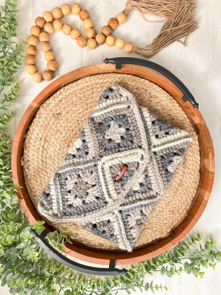 Neutral-tone crochet pouch with granny square pattern on woven tray, greenery, wooden beads, and fringed tan textile nearby.