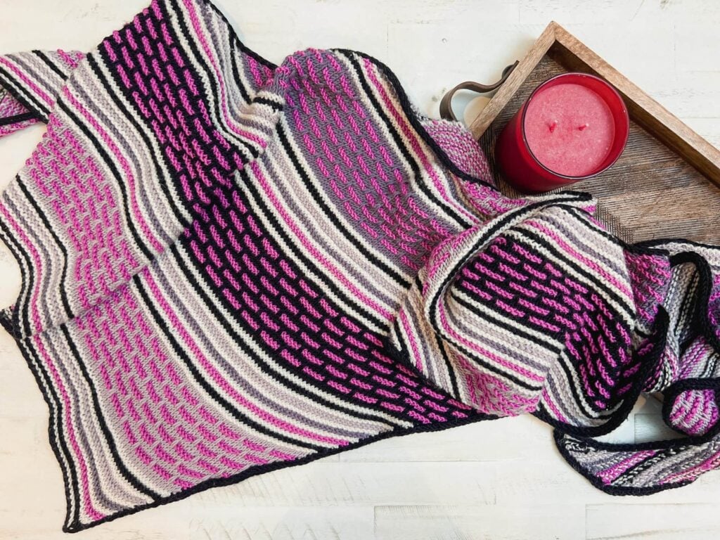 A knitted blanket with black, white, gray, and magenta stripes and patterns—reminiscent of a knit mosaic shawl—is draped on a white surface next to a wooden tray holding a large red candle. -Marly Bird