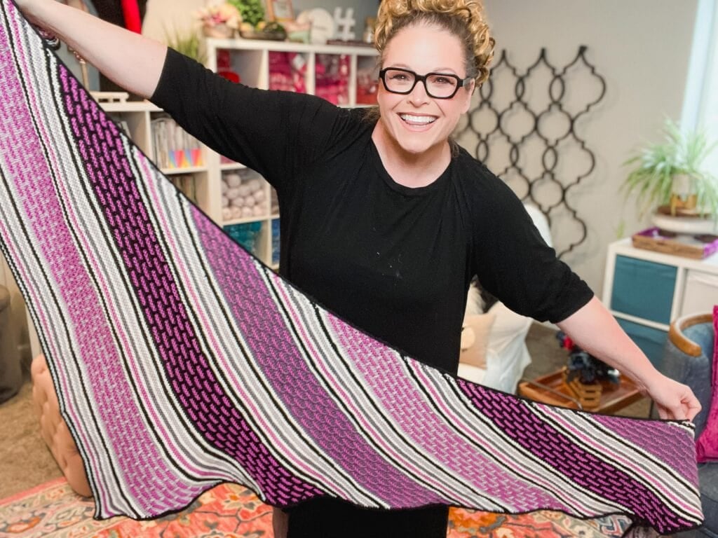 A smiling person wearing glasses and a black dress holds up a large, colorful knit mosaic shawl with pink, purple, black, and white stripes in a cozy, well-decorated craft room. -Marly Bird