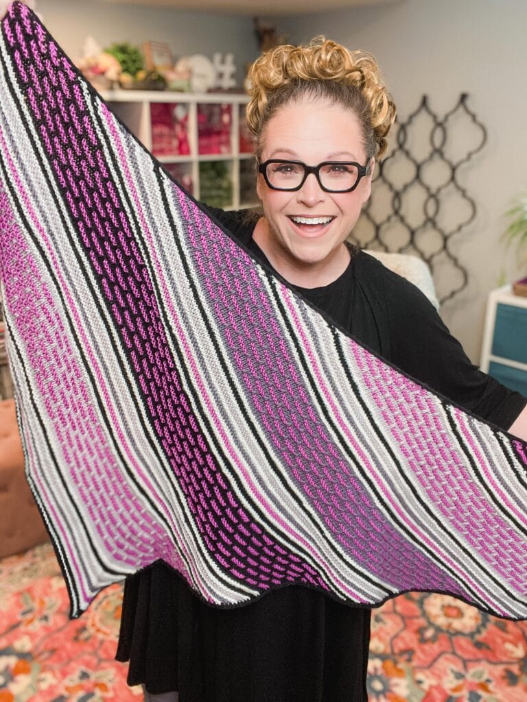 A smiling person with glasses holds up a knitted shawl featuring pink, purple, black, and white stripes with a geometric pattern—an inviting display of box mosaic stitches knitting—in a cozy, well-decorated room. -Marly Bird