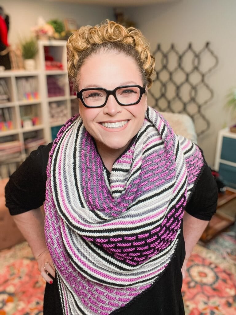 A smiling person with curly blonde hair, black glasses, and a large striped and patterned shawl—made from an easy knit crescent shawl pattern—stands indoors in a cozy, colorfully decorated room. -Marly Bird