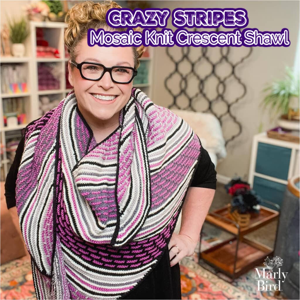A smiling woman wearing glasses shows off a large Knit Crescent Shawl with black, white, purple, and pink stripes. Text reads, "Crazy Stripes Mosaic Knit Crescent Shawl. Marly Bird. -Marly Bird