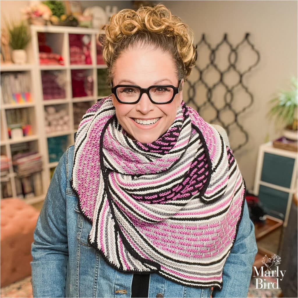 A smiling woman with curly hair and glasses wears a vibrant Knit Crescent Shawl in pink, white, and black stripes over a denim jacket. She stands in a cozy, well-lit room with shelves of yarn in the background. -Marly Bird