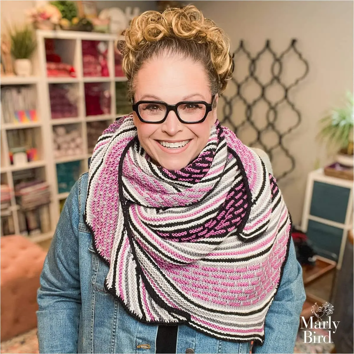 A person models a knit shawl with bold stripes in pink, black, white, and gray; clear stitch texture over a denim jacket.