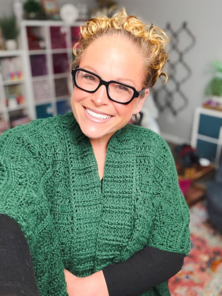 Green crochet ruana shawl with visible openwork stitch, modeled in a bright room with shelves and plants behind.