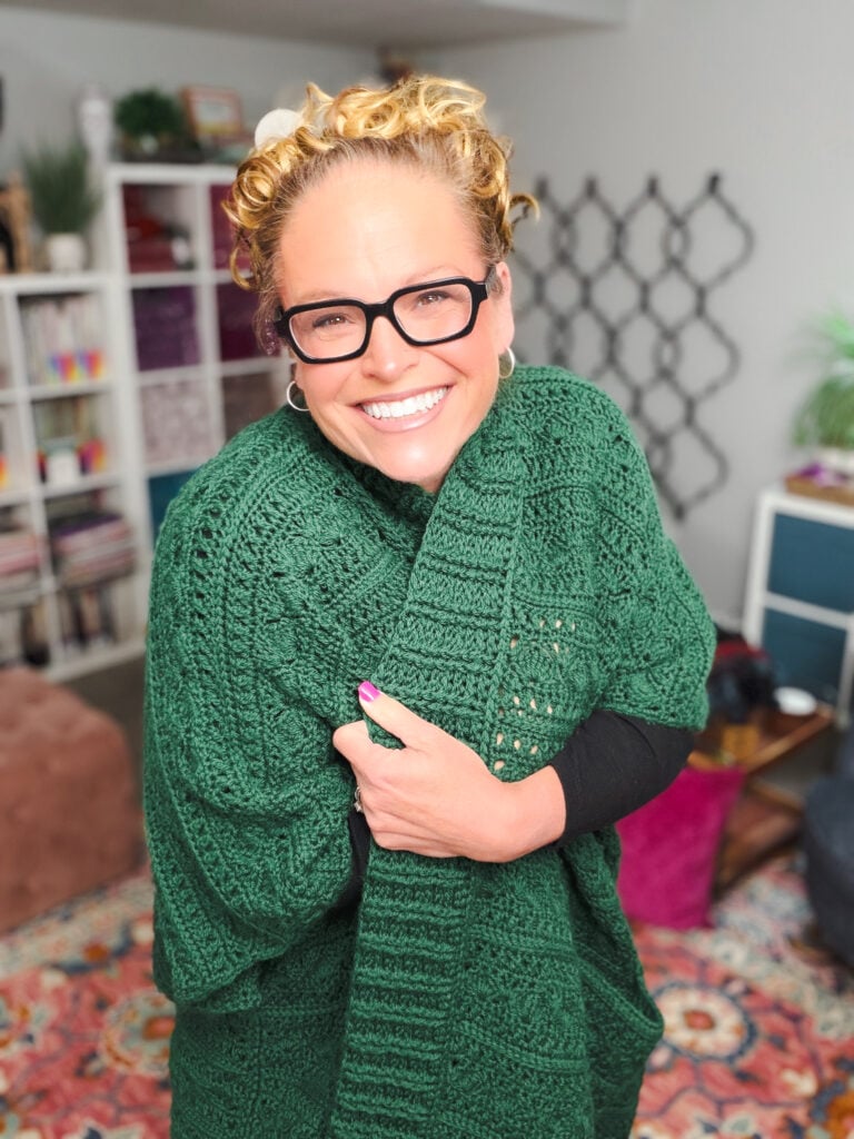 Person models a green crochet ruana, showing textured stitch detail; cozy room with bookshelves and patterned decor in background.