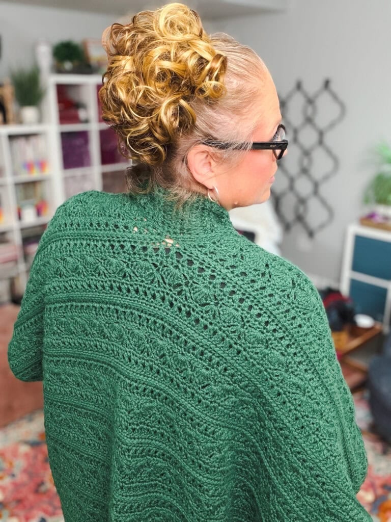 A green crochet ruana shawl with visible stitch patterns is modeled indoors, bookshelves and colorful decor in the background.